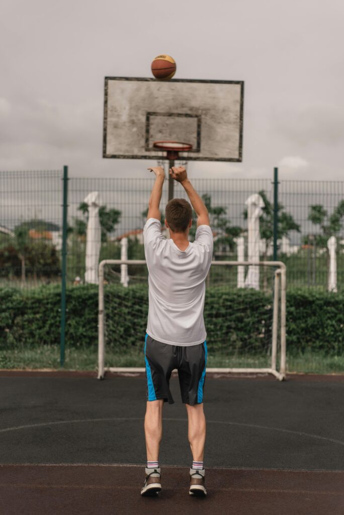 Back view of a young man playing basketball, shooting towards the hoop on an outdoor court.